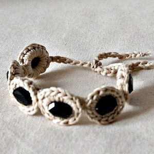 May include: A crocheted bracelet with black beads. The bracelet is made of a natural beige colored string.