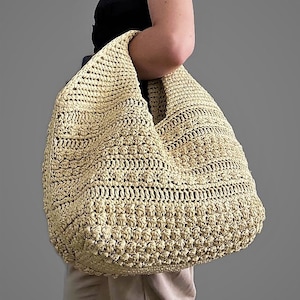 Può includere: Una grande borsa tote in crochet beige chiaro con una forma morbida e non strutturata. La borsa è realizzata con un filato spesso e testurizzato e ha un'ampia apertura superiore.