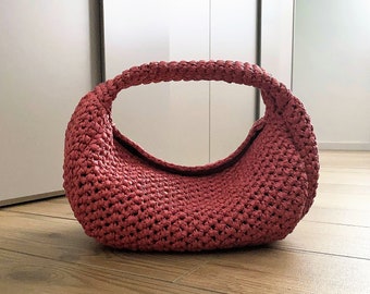 Op de afbeelding: Een gehaakte roze handtas met een afgeronde vorm en een enkele handgreep.