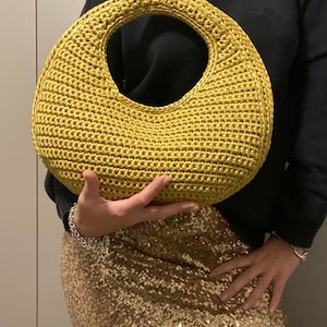 May include: Mustard yellow crocheted handbag with a circular handle. The bag has a rounded shape and is held by a person. The bag is made of a textured yarn. The person is wearing a black sweater and a gold sequined skirt.