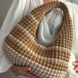 Puede incluir: Un bolso de ganchillo en forma de media luna con un patrón de puntos texturizados. El bolso presenta rayas horizontales en tonos alternos de amarillo mostaza, beige y crema, con un asa gruesa integrada que forma la curva superior.