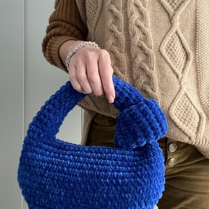 Op de afbeelding: Een blauwe gehaakte handtas met een pluche textuur. De tas heeft een enkele band en een rond, pluche pompon detail.