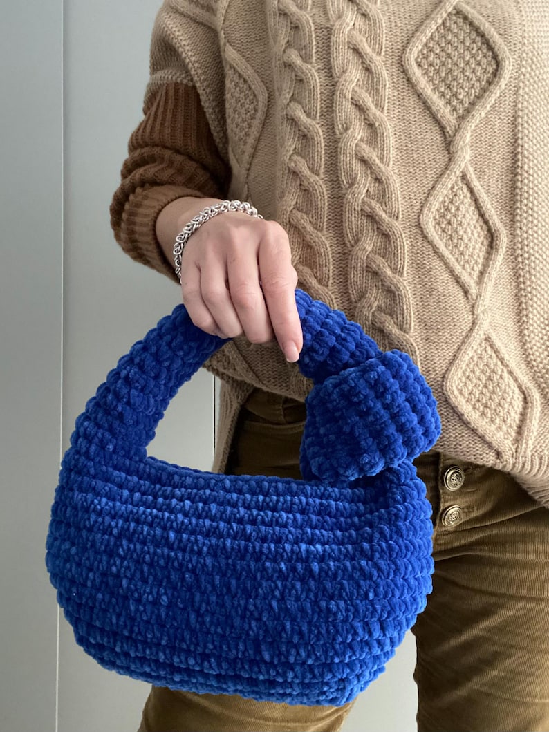 Op de afbeelding: Een blauwe gehaakte handtas met een pluche textuur. De tas heeft een enkele band en een rond, pluche pompon detail.