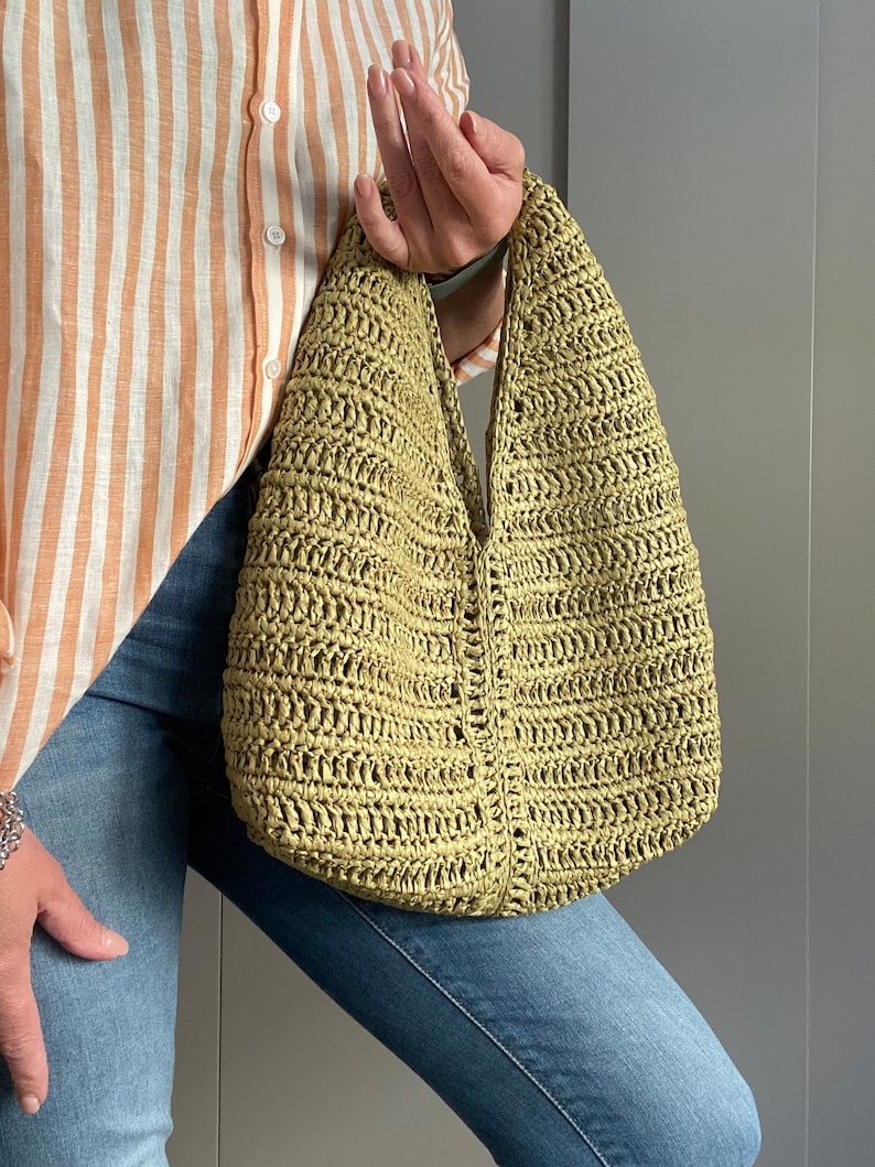 May include: A green crocheted tote bag with a wide, open top and a single strap. The bag is made of a woven material and has a slightly textured surface.