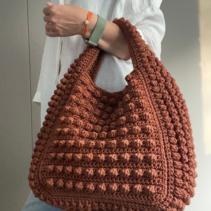 Puede incluir: Un bolso tote grande de crochet marr&oacute;n con una superficie texturizada y abultada. El bolso tiene una parte superior ancha y abierta y dos asas largas.