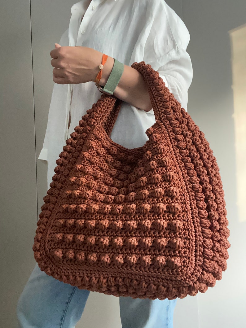 Puede incluir: Un bolso tote grande de crochet marr&oacute;n con una superficie texturizada y abultada. El bolso tiene una parte superior ancha y abierta y dos asas largas.