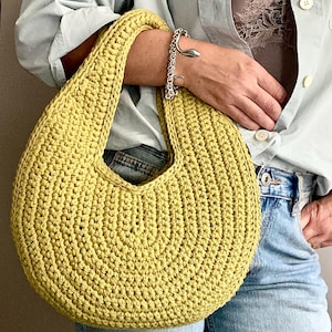 Op de afbeelding: Een geelgroene, gehaakte handtas met een afgeronde vorm en een enkele handgreep. De tas is gemaakt van dik garen en de steken zijn duidelijk zichtbaar. De tas wordt vastgehouden door een persoon die een lichtblauw overhemd en een blauwe spijkerbroek draagt.