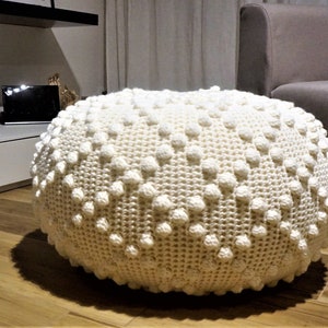 Puede incluir: Un puf redondo de crochet blanco con una superficie texturizada. El puf tiene un diámetro de aproximadamente 46 cm.
