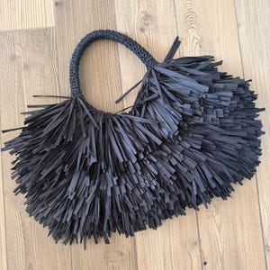 May include: A black handbag featuring a dense, layered fringe design made of thin, flat strips. The bag has a single, rounded, crocheted handle that connects to the main body, which is entirely covered in the long, hanging black fringe material.