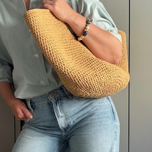 May include: A light brown woven tote bag with a curved shape, carried over the shoulder. The person is wearing a light blue shirt and blue jeans. A bracelet with blue and white beads is visible. The bag is likely made of natural fibres.