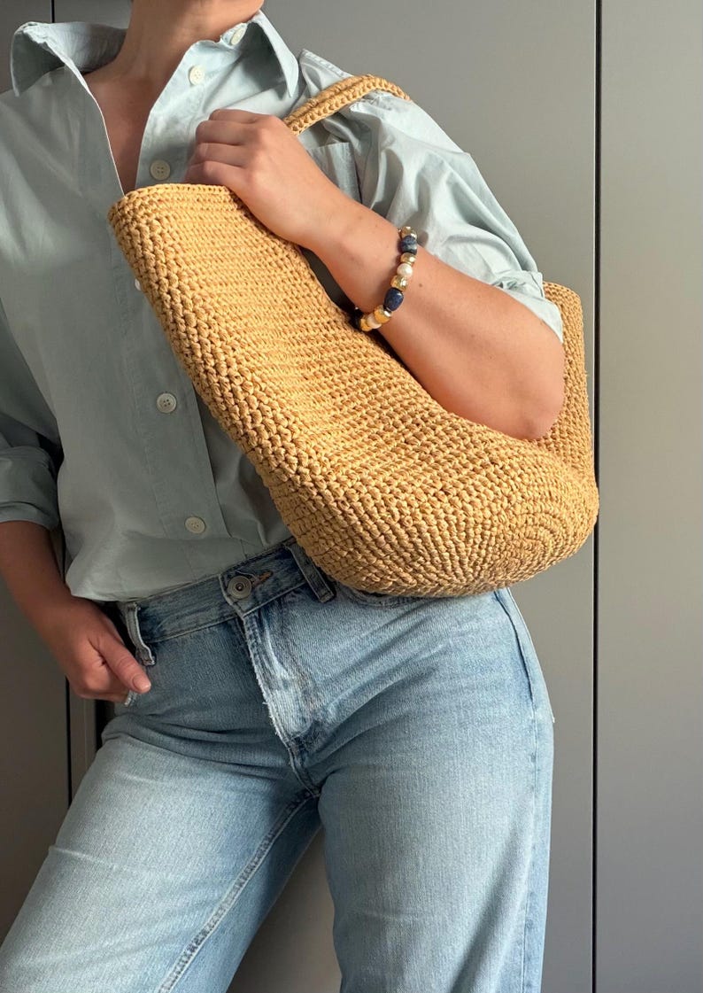 May include: A light brown woven tote bag with a curved shape, carried over the shoulder. The person is wearing a light blue shirt and blue jeans. A bracelet with blue and white beads is visible. The bag is likely made of natural fibres.