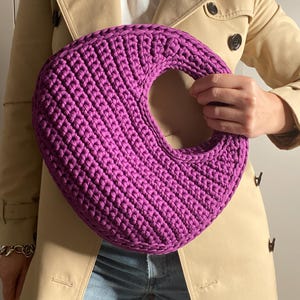 Può includere: Una vivace borsa a mano all'uncinetto color magenta, con un design circolare e una grande apertura centrale. La borsa è presentata contro un trench beige e jeans blu, evidenziando la sua forma e consistenza uniche. Il diametro della borsa è di circa 30 cm.