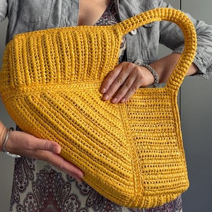 May include: A bright yellow, hand-crocheted tote bag featuring a textured, ribbed stitch pattern. The bag has a structured, angular shape with a single integrated handle, showcasing a consistent, dense weave throughout the entire surface.