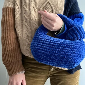 Op de afbeelding: Een blauwe gehaakte handtas met een bruine en beige kabelgebreide trui op de achtergrond.
