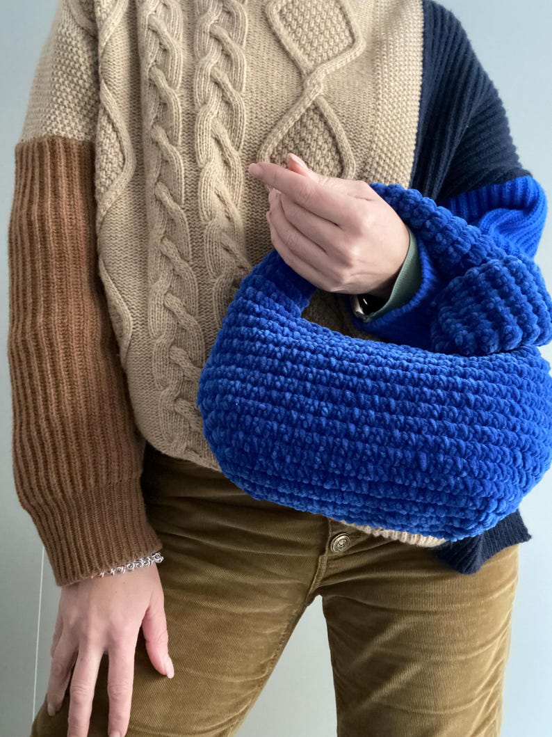 Op de afbeelding: Een blauwe gehaakte handtas met een bruine en beige kabelgebreide trui op de achtergrond.