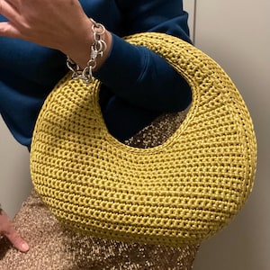 May include: A mustard yellow, crocheted handbag with a large, circular handle. The bag has a rounded shape and a textured surface. The bag is being held by a person wearing a blue sweater and a silver bracelet.