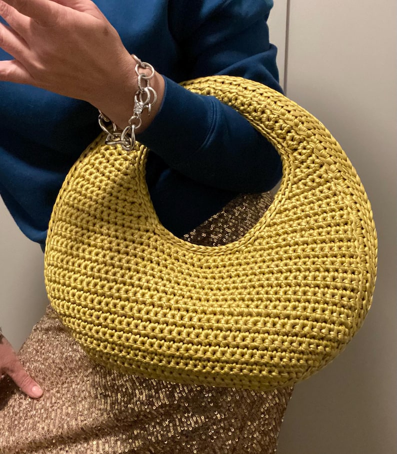 May include: A mustard yellow, crocheted handbag with a large, circular handle. The bag has a rounded shape and a textured surface. The bag is being held by a person wearing a blue sweater and a silver bracelet.