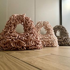 Può includere: Tre borse fatte a mano, strutturate, in varie tonalità di rosa e beige. Ogni borsa ha una forma triangolare con un'apertura circolare e un design ad anello, simile a frange. Le borse sono esposte su un pavimento in legno.