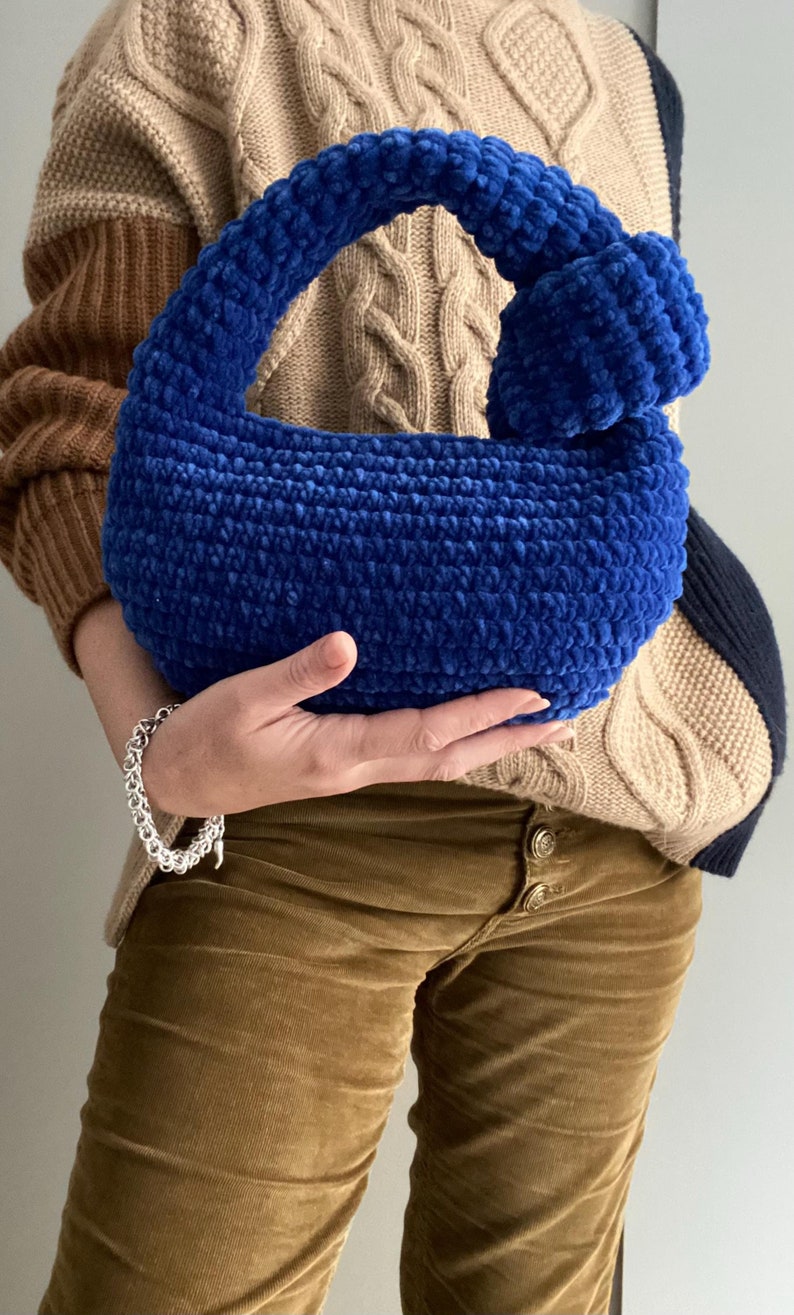 Op de afbeelding: Een blauwe gehaakte handtas met een geknoopte handvat. De tas is gemaakt van zacht, pluizig garen en heeft een afgeronde vorm.