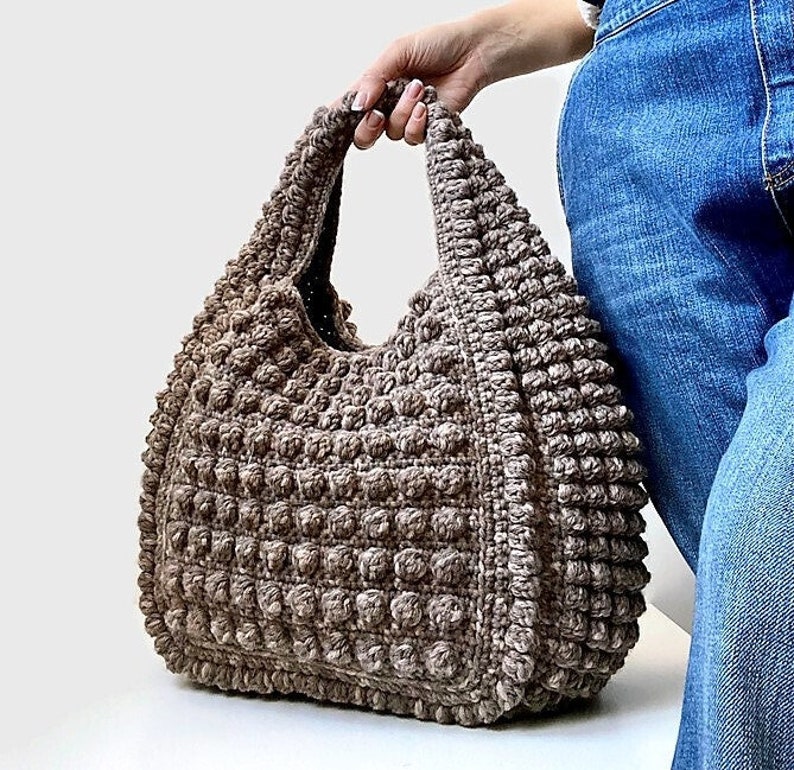 Puede incluir: Un bolso tote de crochet marr&oacute;n con un dise&ntilde;o texturizado y abultado. El bolso tiene una sola correa y es lo suficientemente grande como para llevar los art&iacute;culos esenciales del d&iacute;a a d&iacute;a.