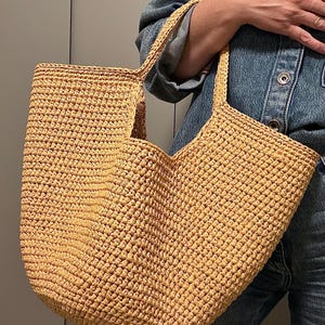 May include: A large, open-top tote bag made of woven, light brown material. The bag has two handles and a rectangular shape. The texture of the material is visible, and the bag appears sturdy. The bag is held by a person wearing a denim jacket.