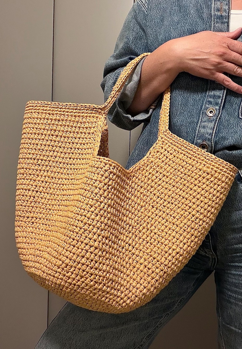 May include: A large, open-top tote bag made of woven, light brown material. The bag has two handles and a rectangular shape. The texture of the material is visible, and the bag appears sturdy. The bag is held by a person wearing a denim jacket.