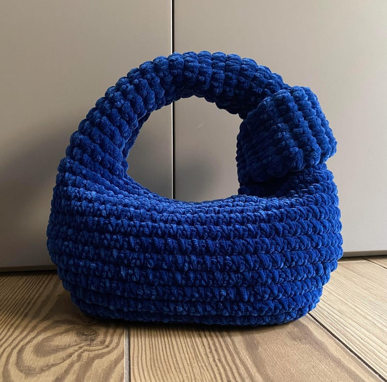 Op de afbeelding: Een blauwe gehaakte handtas met een geknoopte handvat. De tas is gemaakt van zacht, pluche garen.