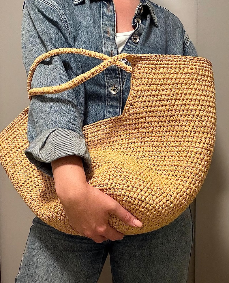 May include: A large, woven straw tote bag in a light tan colour. The bag has a rounded shape with a wide opening and two sturdy handles. The texture of the bag is visible, showing the individual strands of straw. The bag is held by a person.