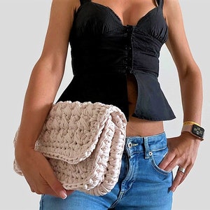 May include: A light pink crocheted clutch bag with a flap closure. The bag is held by a person wearing a black top and blue jeans.
