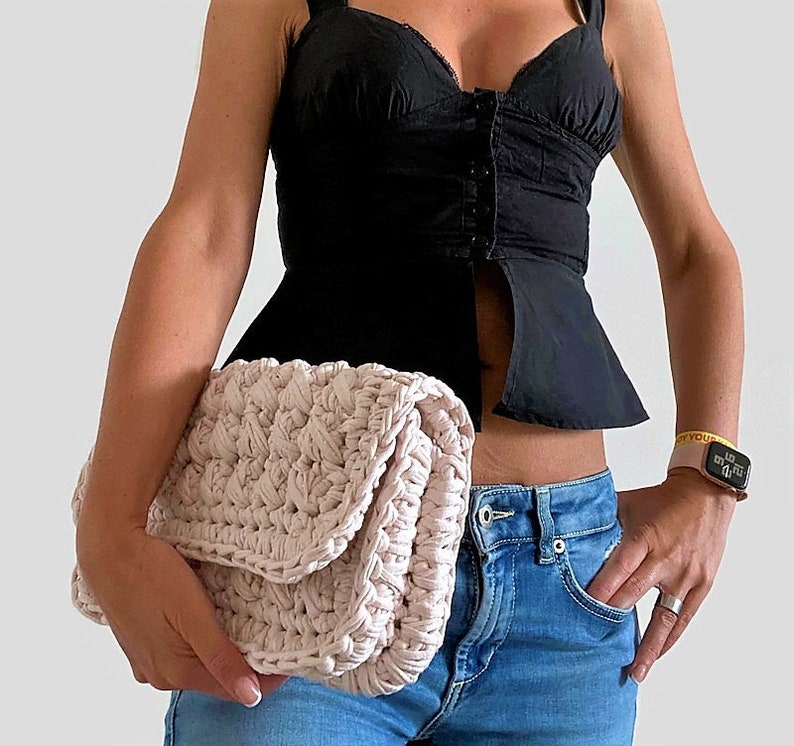 May include: A light pink crocheted clutch bag with a flap closure. The bag is held by a person wearing a black top and blue jeans.
