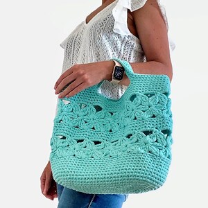May include: A turquoise crocheted tote bag with a flower pattern. The bag has a wide, open top and two handles.