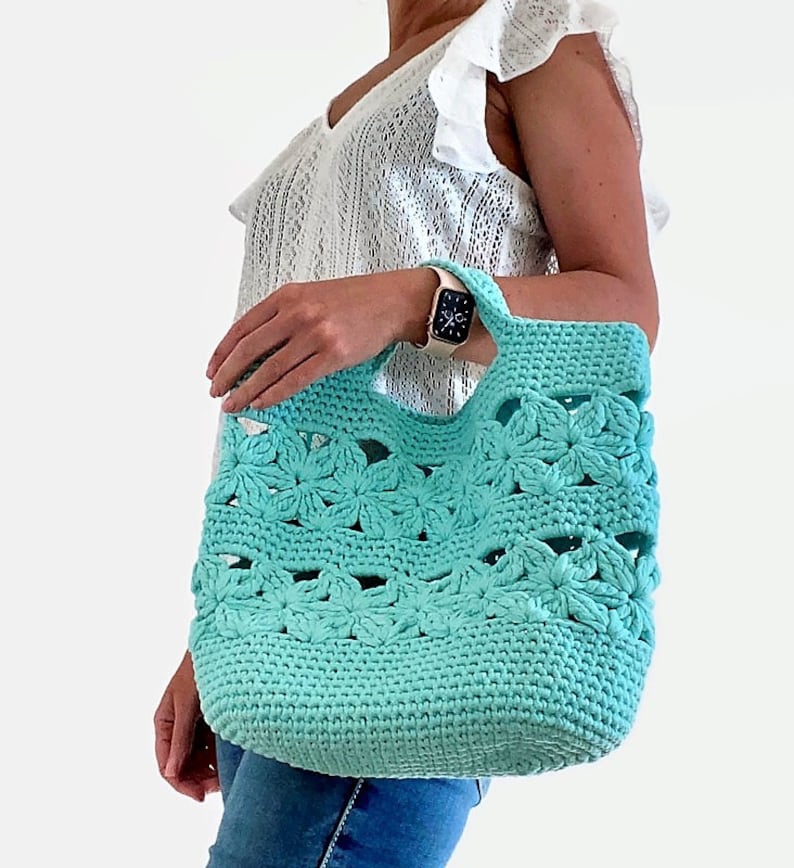May include: A turquoise crocheted tote bag with a flower pattern. The bag has a wide, open top and two handles.