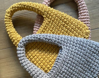 May include: Three crocheted bags in different colors: gray, yellow, and pink. The bags are made with a thick yarn and have a simple, modern design.
