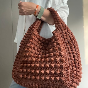 Puede incluir: Un bolso tote grande de crochet marr&oacute;n con una superficie texturizada y abultada. El bolso tiene una parte superior ancha y abierta y dos asas largas.