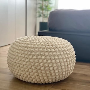 May include: Cream-colored, round ottoman with a textured, bobble-stitch design. The footstool is made of a soft, woven material and sits on a wooden floor. The neutral color and simple design make it a versatile piece of furniture.