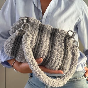 May include: A textured gray crochet handbag featuring alternating stripes of fuzzy light gray yarn and smooth dark gray yarn. The bag has a rounded shape, a matching fuzzy crochet shoulder strap, and large silver-toned metal rings connecting the strap.