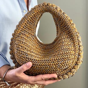 May include: A crescent-shaped handbag crafted from a textured, braided cord in shades of metallic gold and muted grey. The bag features a continuous, thick, rounded border edge and a singular integrated handle opening at the top centre.