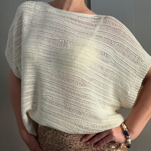 Puede incluir: Top de crochet color crema con tejido abierto, cuello barco y mangas cortas. El top tiene un ajuste holgado y fluido y un aspecto texturizado. El patrón de crochet crea un sutil efecto de rayas. El top se combina con una falda dorada con lentejuelas.