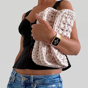 May include: A pink crocheted bag with a textured design. The bag is being held by a person wearing a black top and blue jeans.