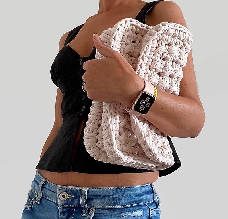 May include: A pink crocheted bag with a textured design. The bag is being held by a person wearing a black top and blue jeans.