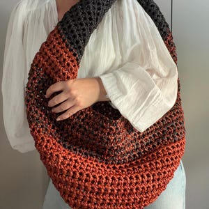 Puede incluir: Un bolso hobo grande en forma de media luna, tejido con hilo grueso en color gris carbón oscuro y naranja quemado con un punto de ganchillo voluminoso. El bolso cuenta con una correa ancha integrada y una silueta redondeada y holgada.
