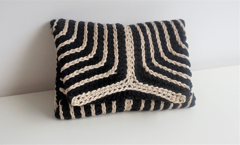 Puede incluir: Un embrague tejido negro y beige con un patr&oacute;n geom&eacute;trico. El embrague est&aacute; hecho de una tela suave y texturizada y tiene un dise&ntilde;o simple y elegante.