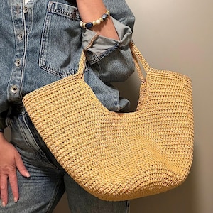 May include: A woven straw tote bag in a light tan hue, featuring a curved top and two handles. The bag has a textured surface. It is held by someone wearing a denim jacket and jeans.