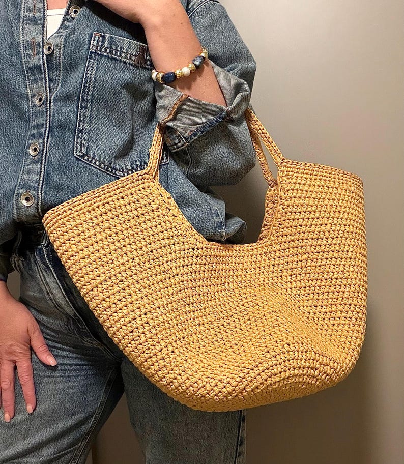 May include: A woven straw tote bag in a light tan hue, featuring a curved top and two handles. The bag has a textured surface. It is held by someone wearing a denim jacket and jeans.