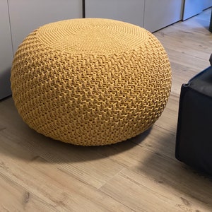 May include: A round, tan crocheted ottoman with a woven pattern. The ottoman is sitting on a light brown wood floor.