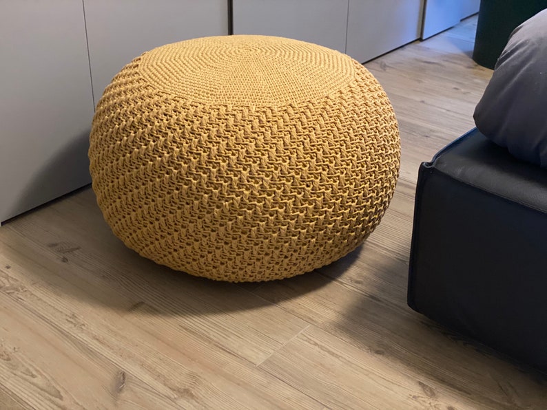 May include: A round, tan crocheted ottoman with a woven pattern. The ottoman is sitting on a light brown wood floor.