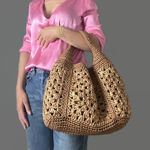 Puede incluir: Un bolso tote grande de color tostado, tejido a mano en crochet, con un patrón abierto de estrellas repetitivas en todo el cuerpo. El bolso tiene una correa de hombro ancha integrada y una silueta estructurada y holgada de material texturizado.