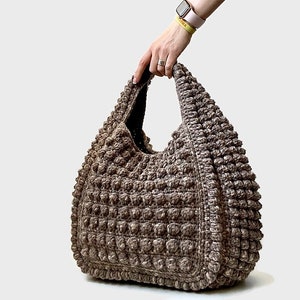 Puede incluir: Un bolso tote de crochet marr&oacute;n con una superficie texturizada y abultada. El bolso tiene un solo asa y una parte superior ancha y abierta.