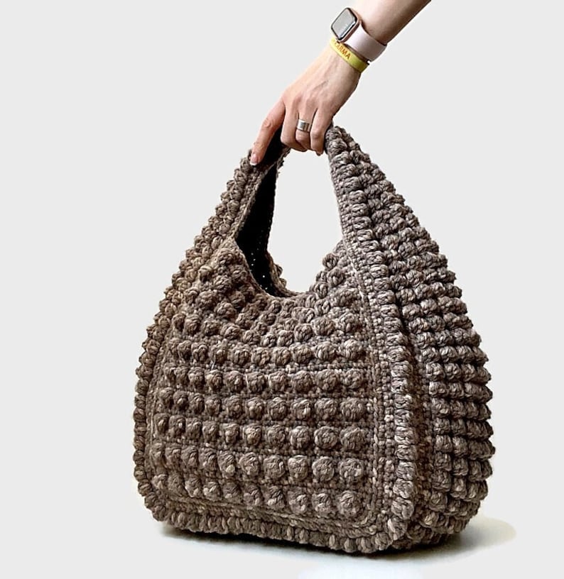 Puede incluir: Un bolso tote de crochet marr&oacute;n con una superficie texturizada y abultada. El bolso tiene un solo asa y una parte superior ancha y abierta.