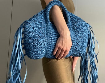 May include: A blue crocheted tote bag with long fringe. The bag has a woven texture and is held by a person.